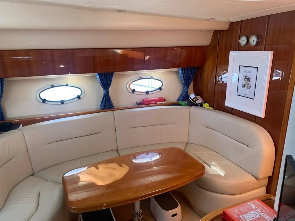 Interior of a boat showcasing a curved leather sofa, wooden table, nautical decor, and round windows with blue curtains.